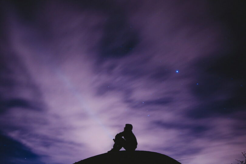 Image of a solitary person sitting on a hill at night beneath a purple sky who yearns for spiritual connection